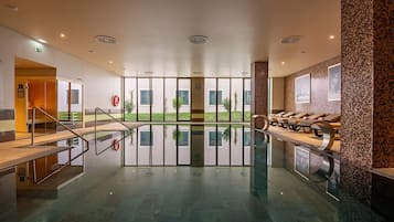 Una piscina interior moderna con paredes de vidrio transparente, una terraza con sol y sillones de descanso.