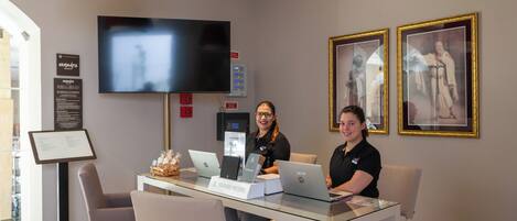 A reception area with a flat-screen TV, framed pictures on the wall, and two people at a desk with laptops.