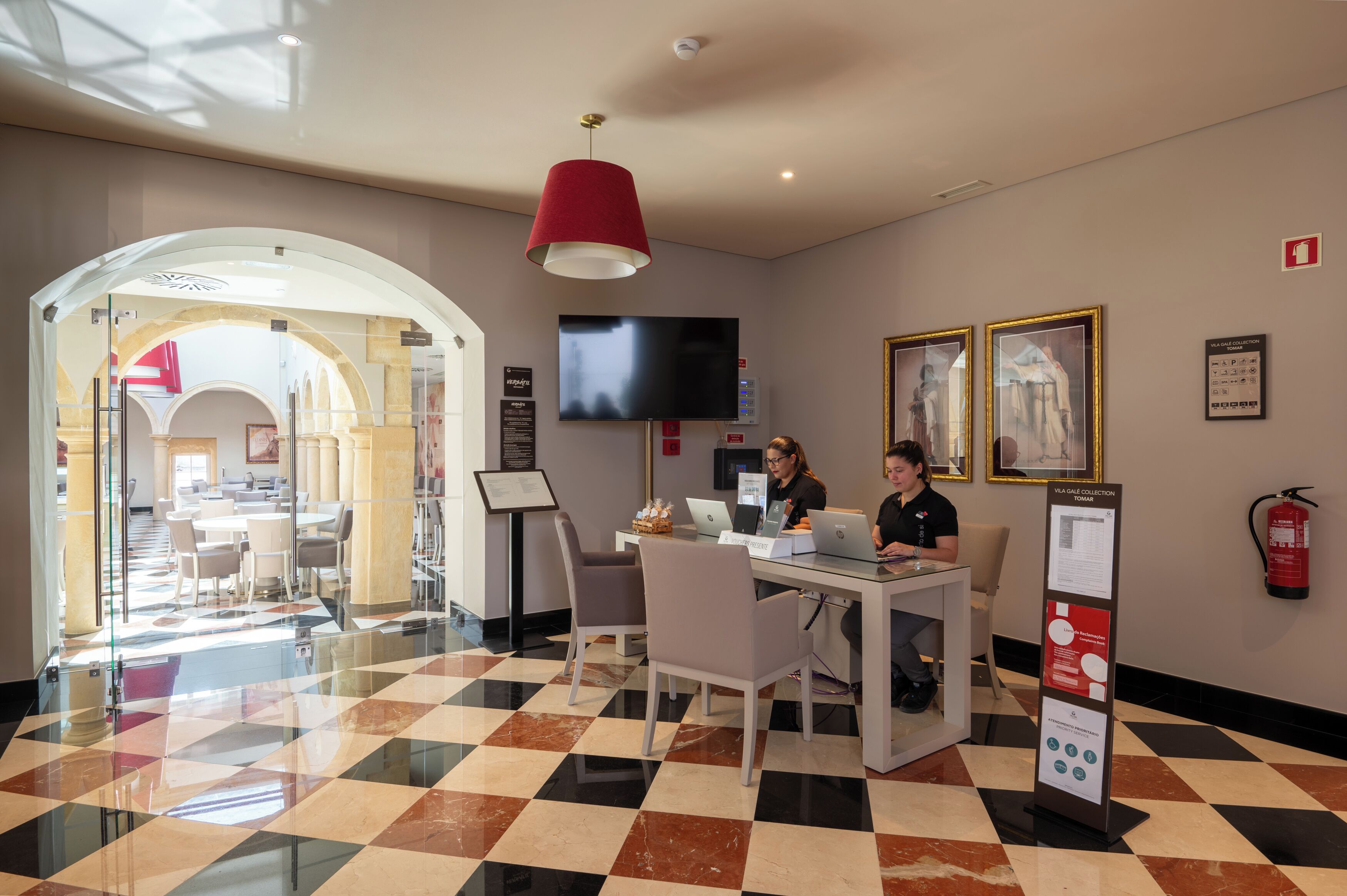 A hotel lobby with a checkered floor, a glass partition, a red pendant light, a flat-screen TV, and a fire extinguisher on the wall.