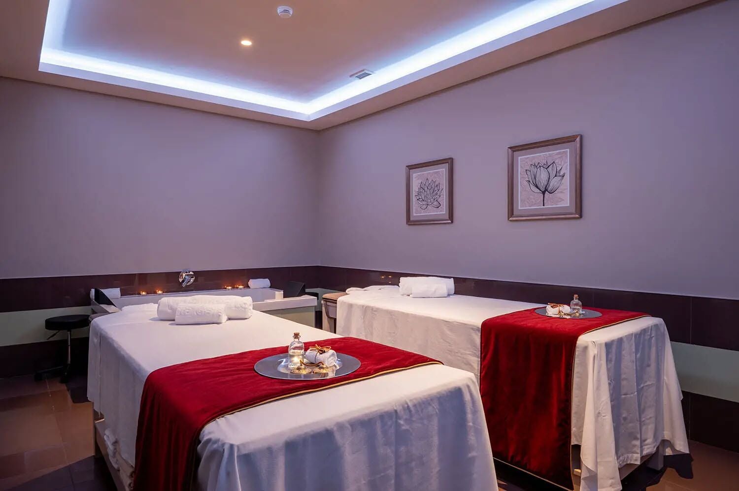 A spa room with two massage tables, each covered with a white sheet and a red blanket. There are framed artworks on the wall, and a small table with a bottle and a towel on each massage table.