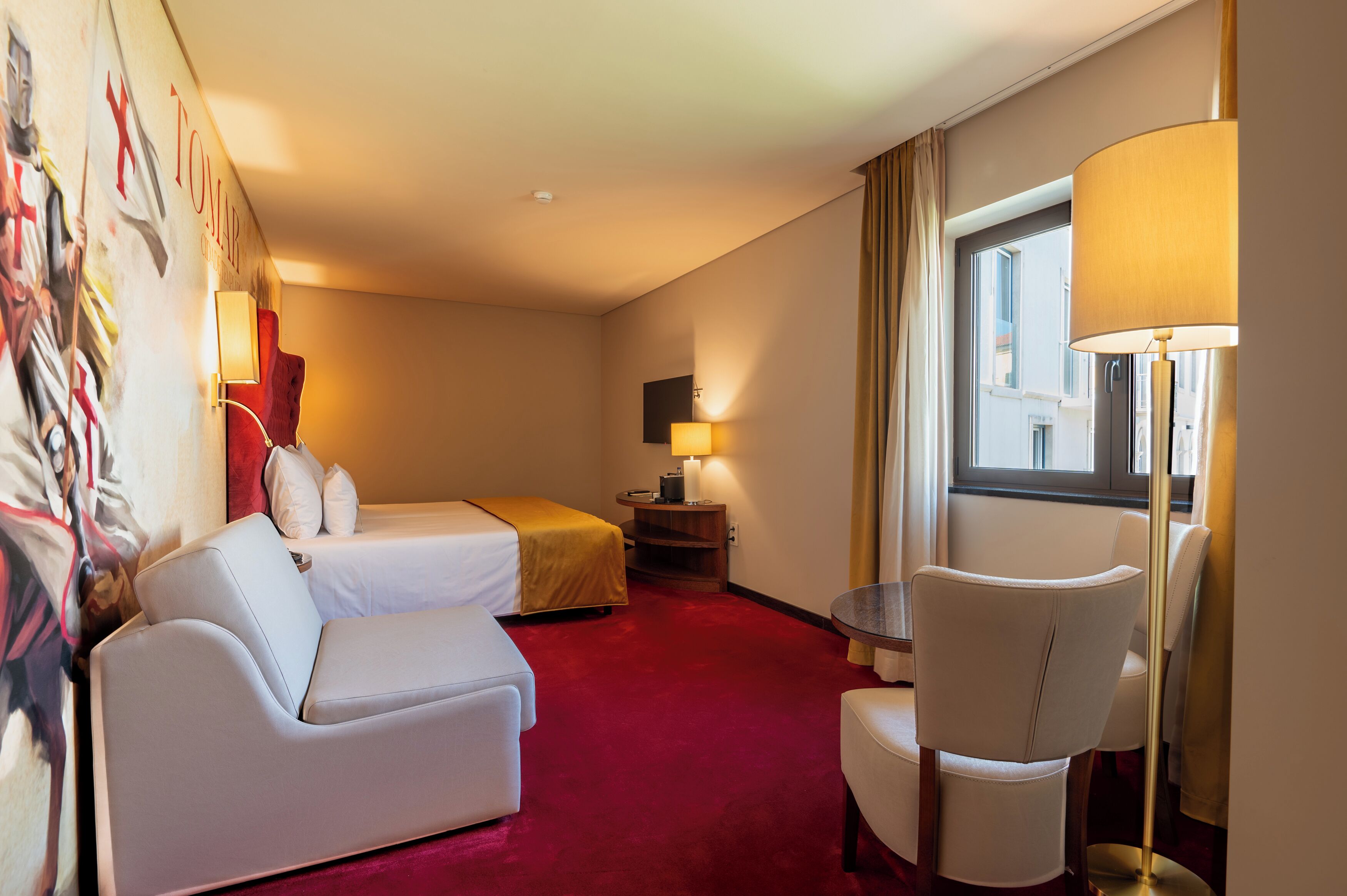A hotel room with a bed, a sofa, a chair, a table, a lamp, and a painting on the wall.