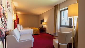 A hotel room with a bed, a sofa, a chair, a table, a lamp, and a painting on the wall.