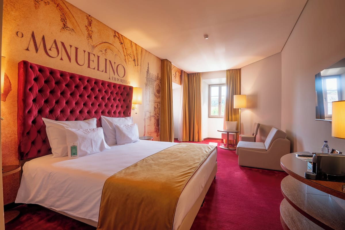 Quarto de hotel com uma grande cabeceira estofada, cama com roupa de cama branca, tapete vermelho, escrivaninha com luminária e uma cadeira.