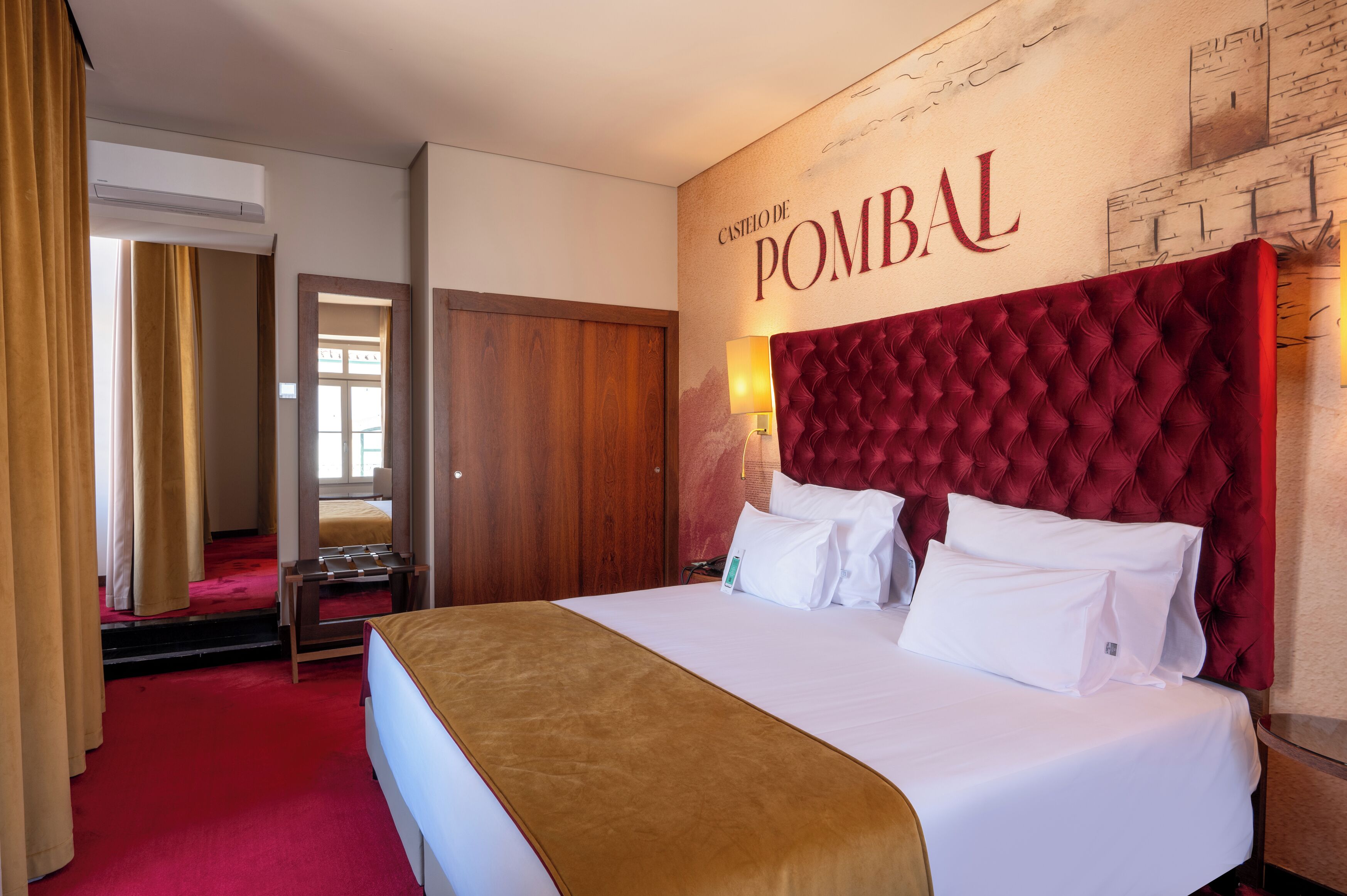 Une chambre d’hôtel avec un grand lit, une tête de lit rouge, un miroir, une armoire en bois et une moquette rouge.