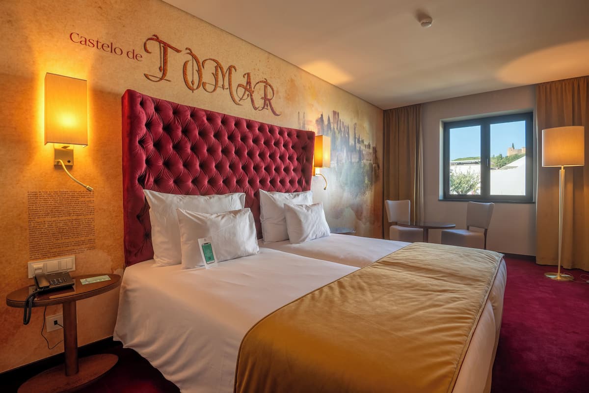 Quarto de hotel com uma cama grande, cabeceira estofada vermelha, luminária na parede, mesa de cabeceira com telefone e janela com vista para um castelo.