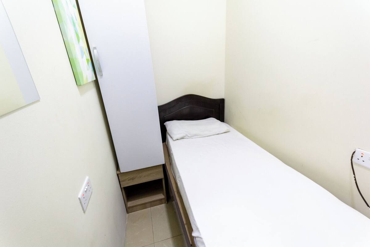 A small, single-bed room with a wooden headboard, a white mattress, and a small shelf under the bed. There is a white wall socket and a door with a green and white door handle.