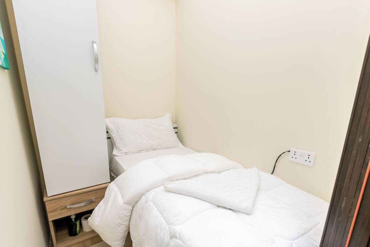 A small, neatly made bed with white bedding in a room with a wooden wardrobe and a power outlet on the wall.