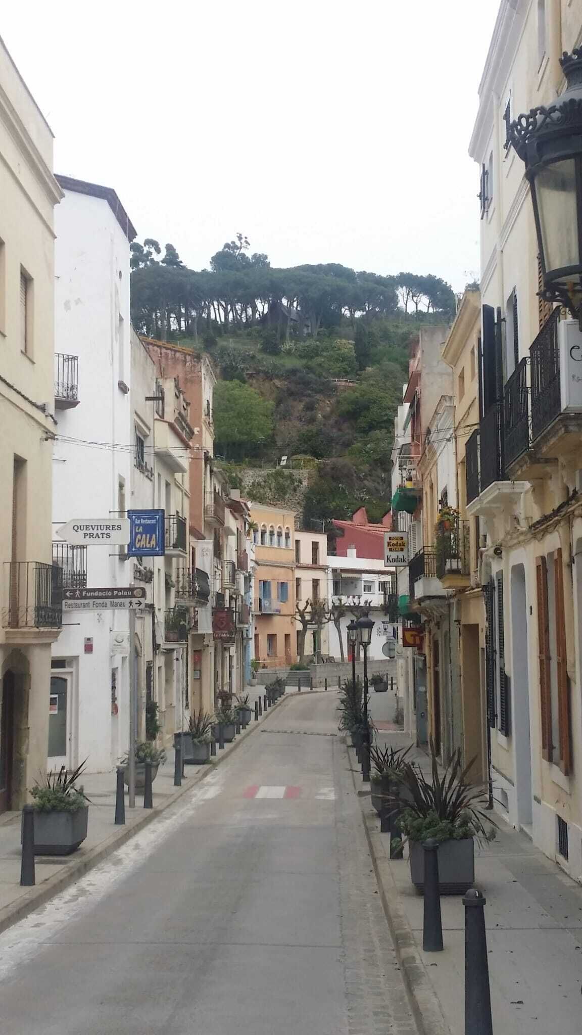 Eine enge Straße, gesäumt von Häusern, von denen einige Balkone und Kübelpflanzen haben. Schilder weisen auf 'QUEVIRES PRATS' und das Restaurant 'LA CALA' hin. Die Straße führt einen Hügel hinauf, im Hintergrund sind Bäume zu sehen.