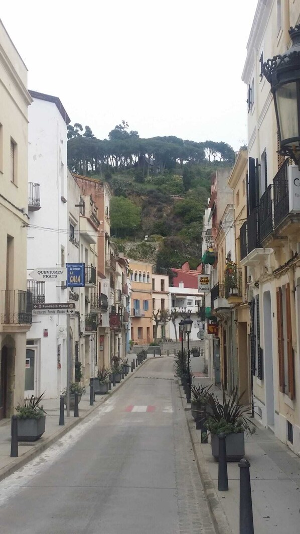 Una calle estrecha bordeada de edificios, algunos con balcones y plantas en macetas. Se ven letreros que indican 'QUEVIRES PRATS' y el restaurante 'LA CALA'. La calle sube una colina con árboles al fondo.