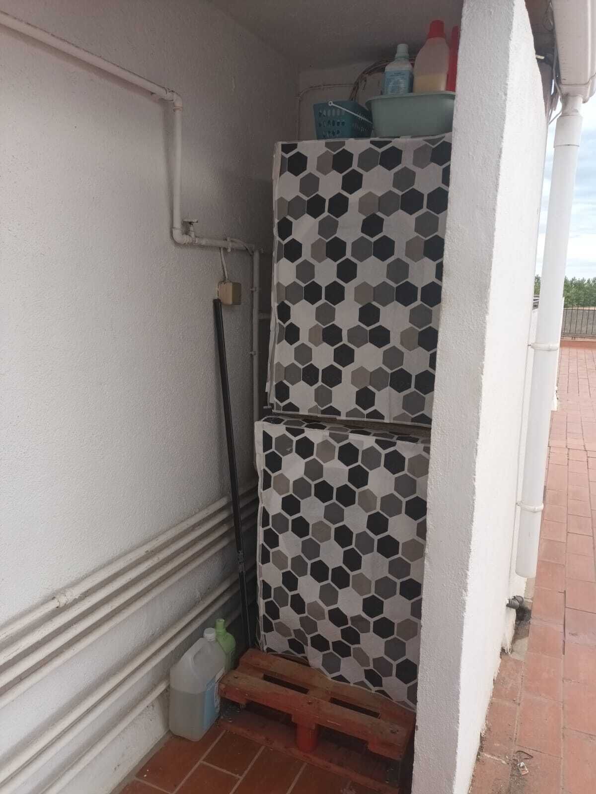 A small, enclosed outdoor area with a tiled wall featuring a geometric pattern. There is a wooden pallet on the ground, a white pipe on the wall, and a small container on the floor.