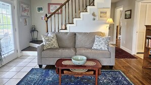 A living room with a grey sofa, a wooden coffee table, and a blue patterned rug. A staircase with wooden railings leads to another level.