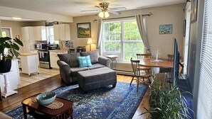 A spacious living room with a grey sofa, a coffee table, and a dining area with a wooden table and chairs. There is a kitchen in the background with white cabinets and a microwave.