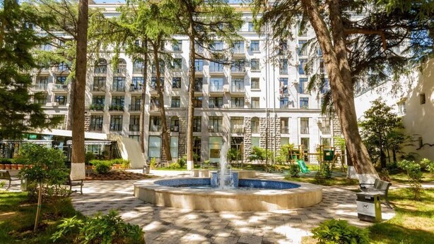 Un patio con una fuente, rodeado de árboles y un edificio de varios pisos con balcones.