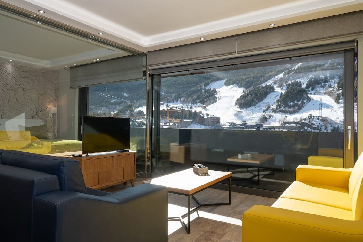 A modern living room with a large TV, yellow and blue sofas, and a view of a snowy mountain landscape.