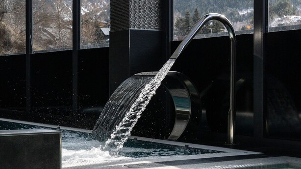 Piscina interior moderna con caída de agua, ventanas amplias que ofrecen vistas a las montañas y suelo de baldosas.