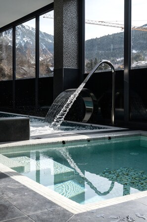Piscina interior moderna com cascata, grandes janelas com vista para as montanhas e piso ladrilhado.