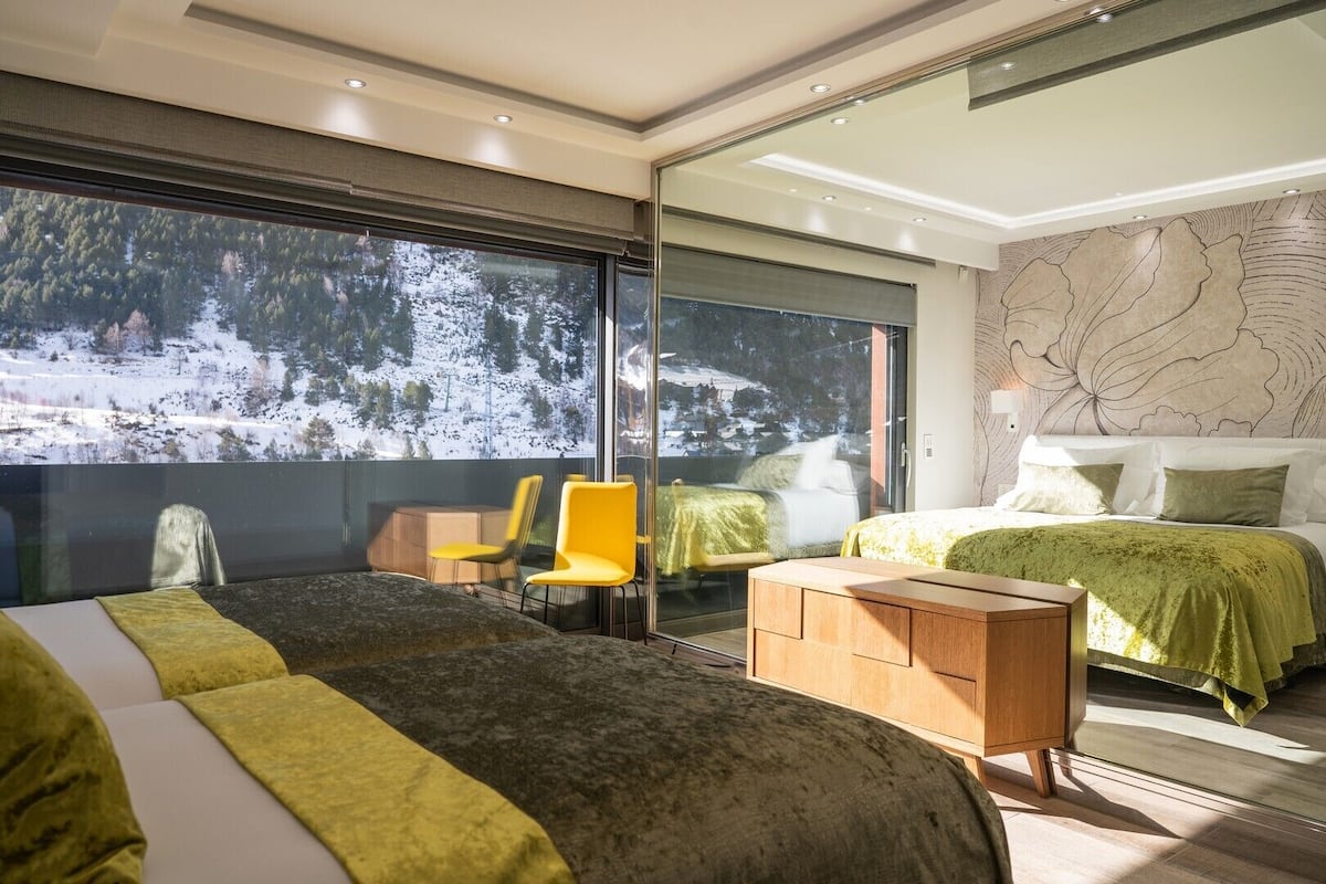 A modern hotel room with a large bed, a small desk, a chair, and a view of a snowy landscape through large windows.