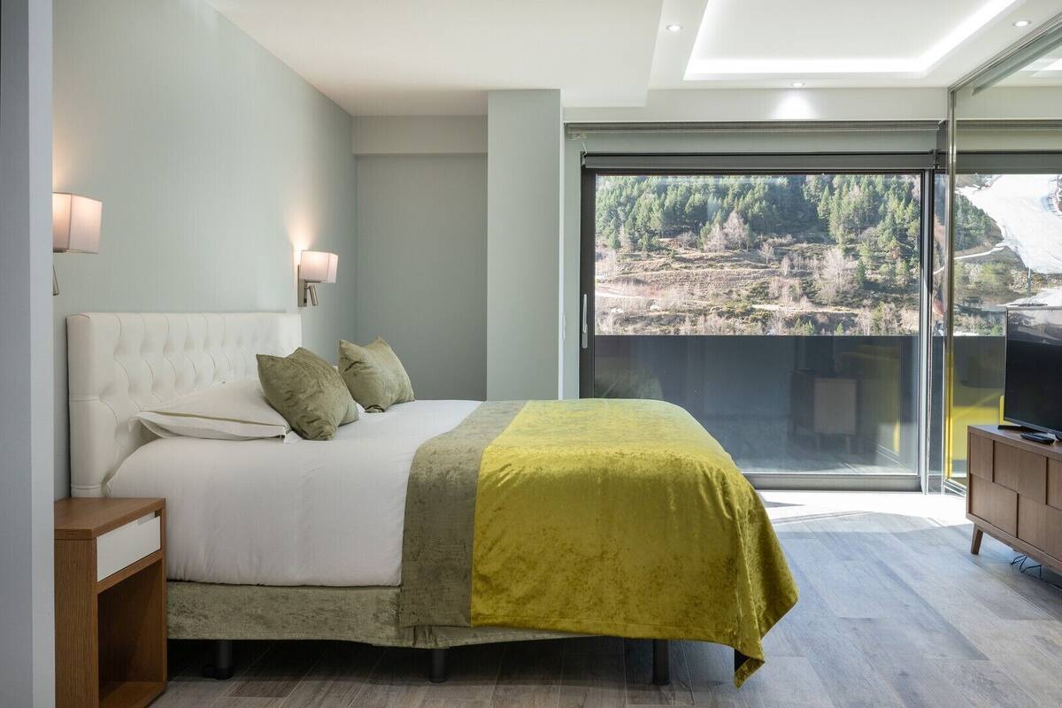 A modern bedroom with a large bed, a TV on a wooden cabinet, and a balcony with a scenic view.