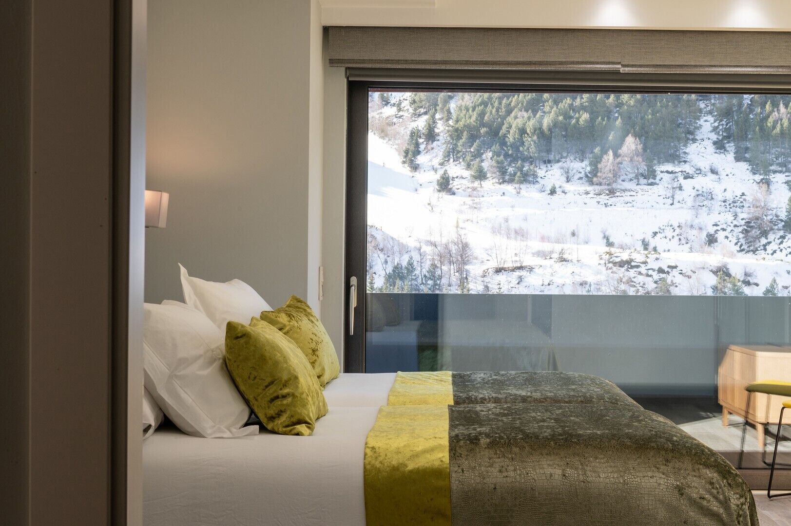 Un dormitorio con cama, almohadas, vistas de un paisaje nevado a través de una ventana y un escritorio de madera con una silla.