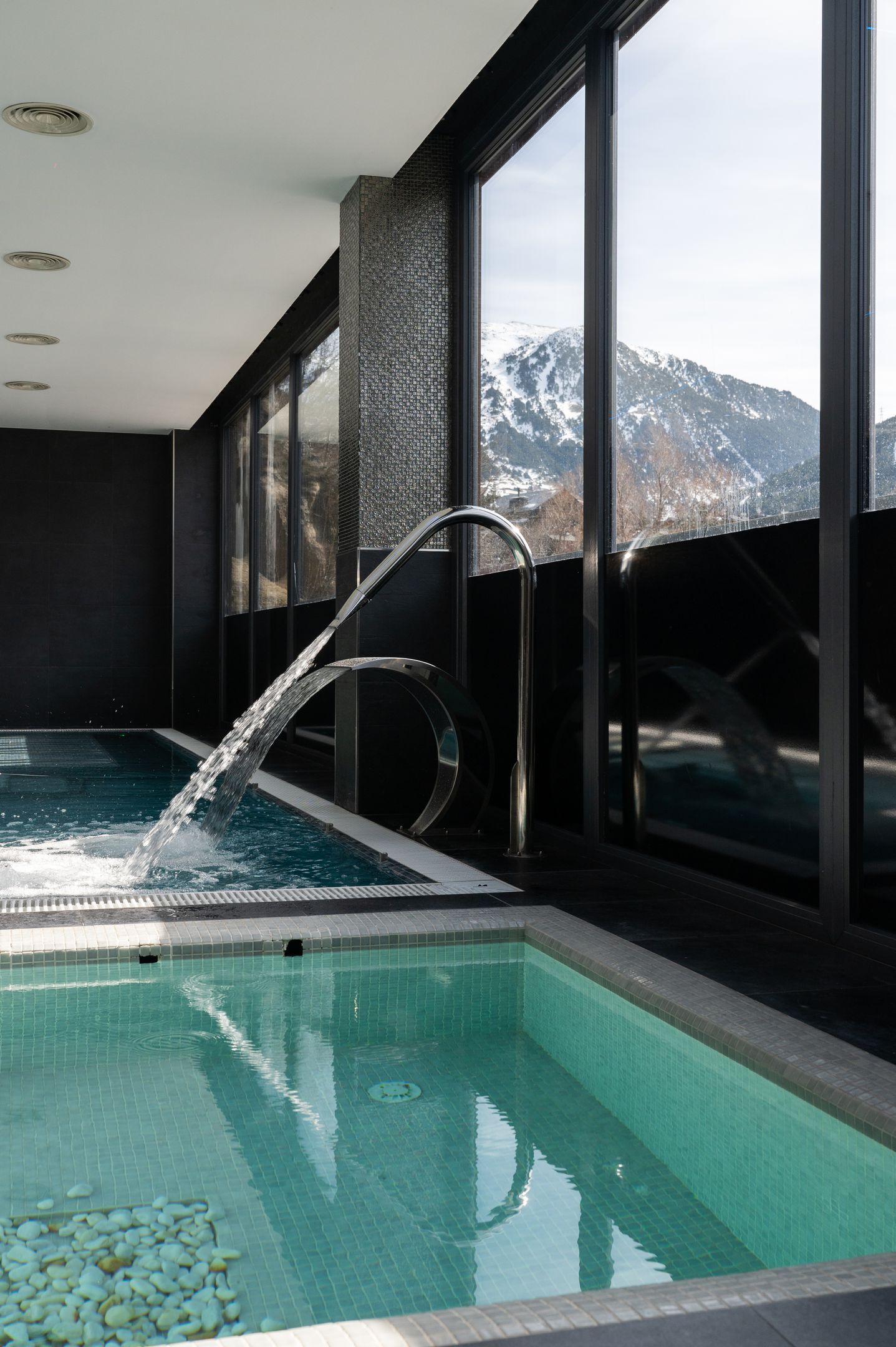 A modern indoor swimming pool with a waterfall feature, large windows offering a view of snow-capped mountains, and a tiled floor.