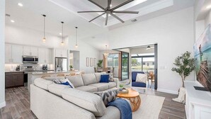 Un salon moderne doté d’un grand canapé d’angle, d’un coin repas avec une table et des chaises, d’une cuisine avec des meubles blancs et des appareils électroménagers, et d’un ventilateur de plafond.