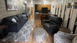 Een ruimtelijk ingerichte leefruimte met een bank, een voetenbankje, een leren fauteuil, houten vloeren, en een keuken met houten kastjes en een magnetron.