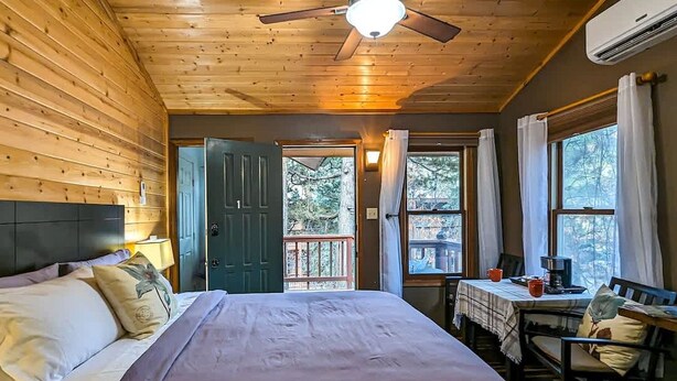 A wooden cabin room with a bed, a desk, and a window overlooking a deck. - Cottages at Canyon Creek #1 (Ruidoso)