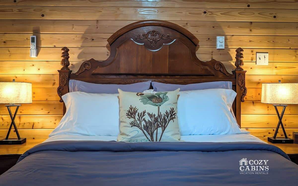 Een houten bed met een gebeeldhouwd hoofdbord, wit beddengoed en een bloemenkussen. Twee nachtlampjes en een digitale klok hangen aan de muur.