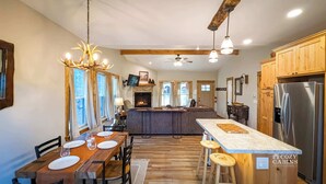 L’intérieur chaleureux d’un chalet avec une table à manger en bois, des tabourets de bar, une cheminée et une cuisine moderne équipée d’un réfrigérateur en acier inoxydable.