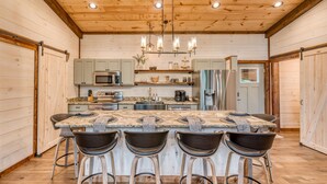 A kitchen with a central island, bar stools, wooden cabinets, and a hanging light fixture. - EPIC Heated pool, Outdoor Fireplace, Pool & Hot Tub! Arcade, and Barrel Sauna (Mineral Bluff)