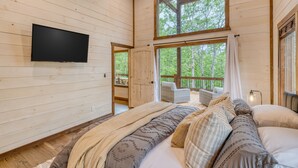 A cozy bedroom with a large bed, a flat-screen TV, and a balcony with a view of the forest. - EPIC Heated pool, Outdoor Fireplace, Pool & Hot Tub! Arcade, and Barrel Sauna (Mineral Bluff)