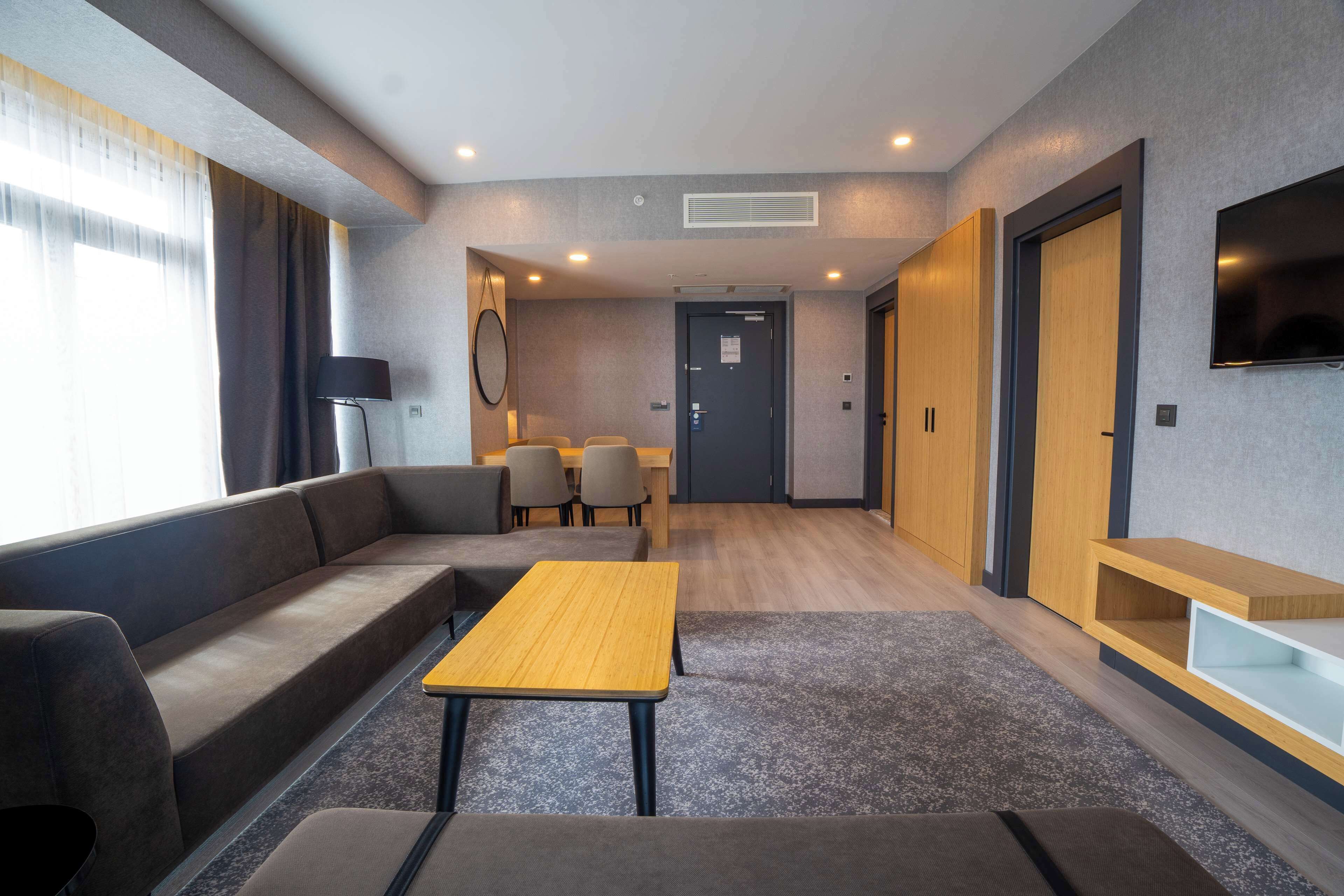 Quarto de hotel moderno com sofá, uma mesa de madeira e televisão fixada na parede.