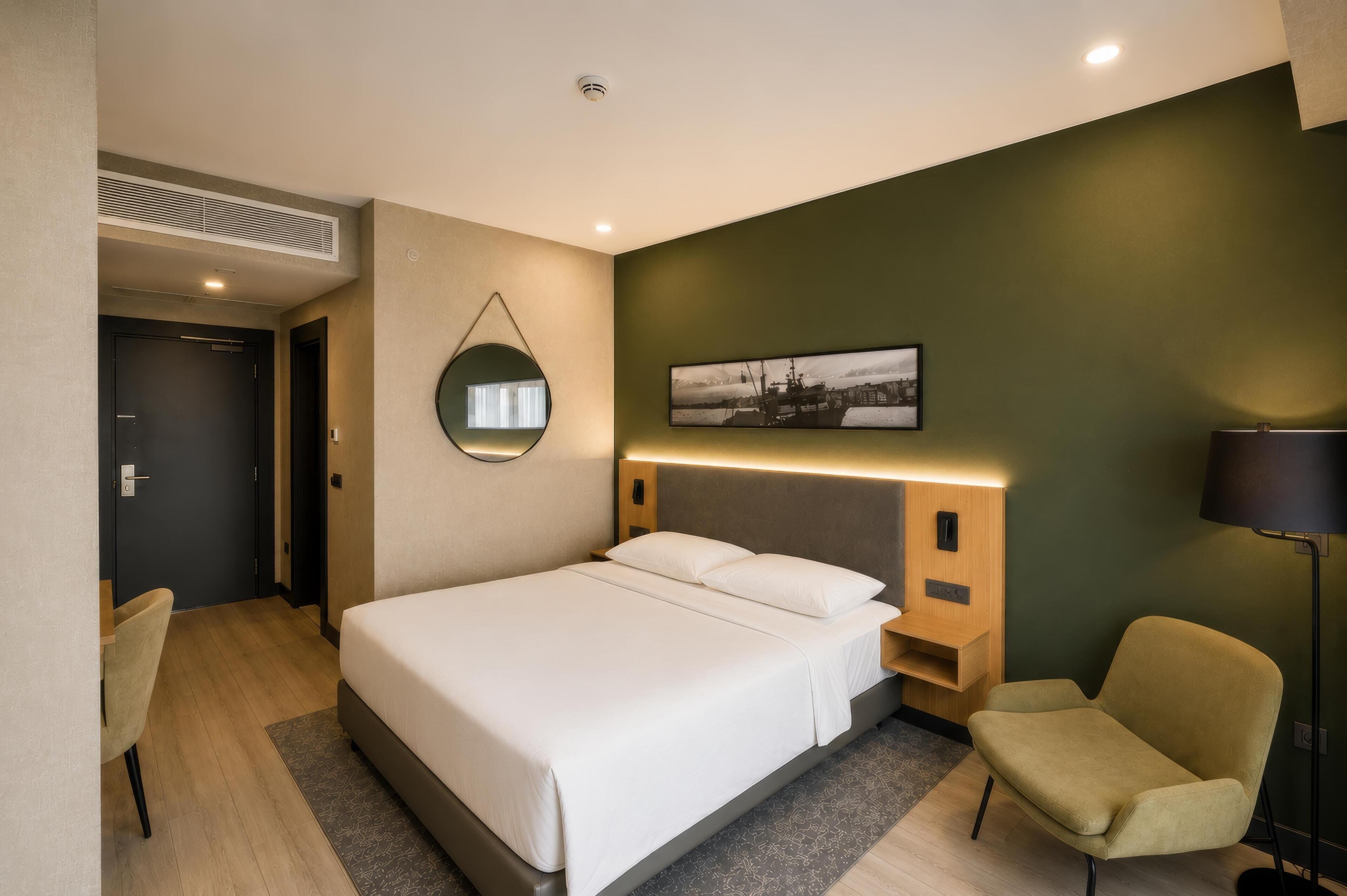 Habitación de hotel con cama, mesita de noche, silla y espejo fijado a la pared.
