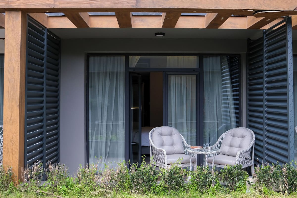 Een balkon met twee rieten stoelen en een tafel, een houten pergola erboven en uitzicht op een kamer met een bed.