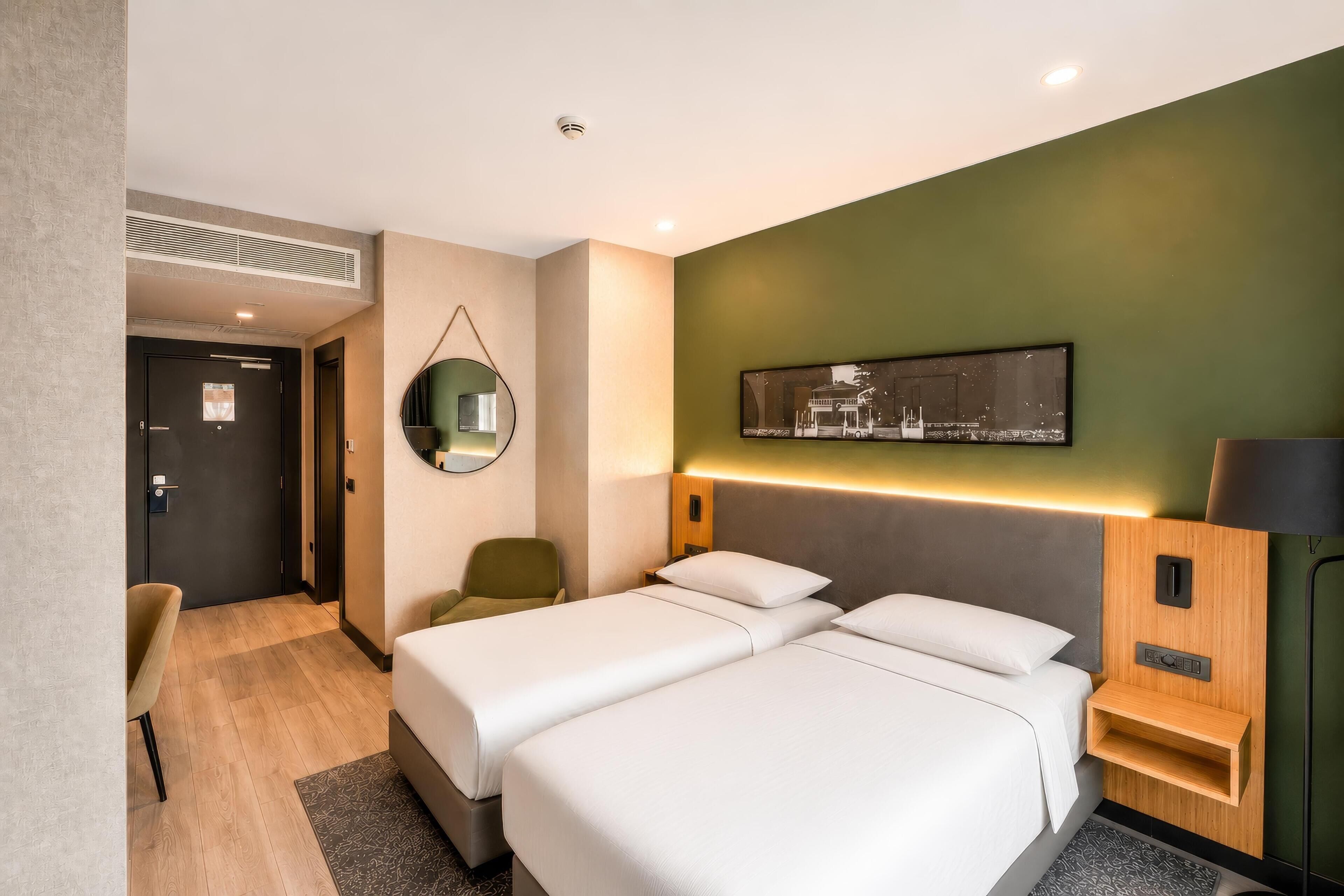 A hotel room with two beds, a desk, a chair, a mirror, a wall-mounted TV, and a bedside table with a lamp.