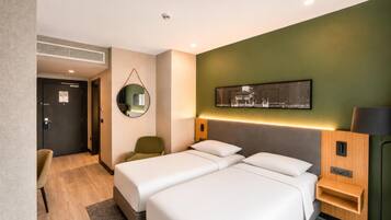 A hotel room with two beds, a desk, a chair, a mirror, a wall-mounted TV, and a bedside table with a lamp.