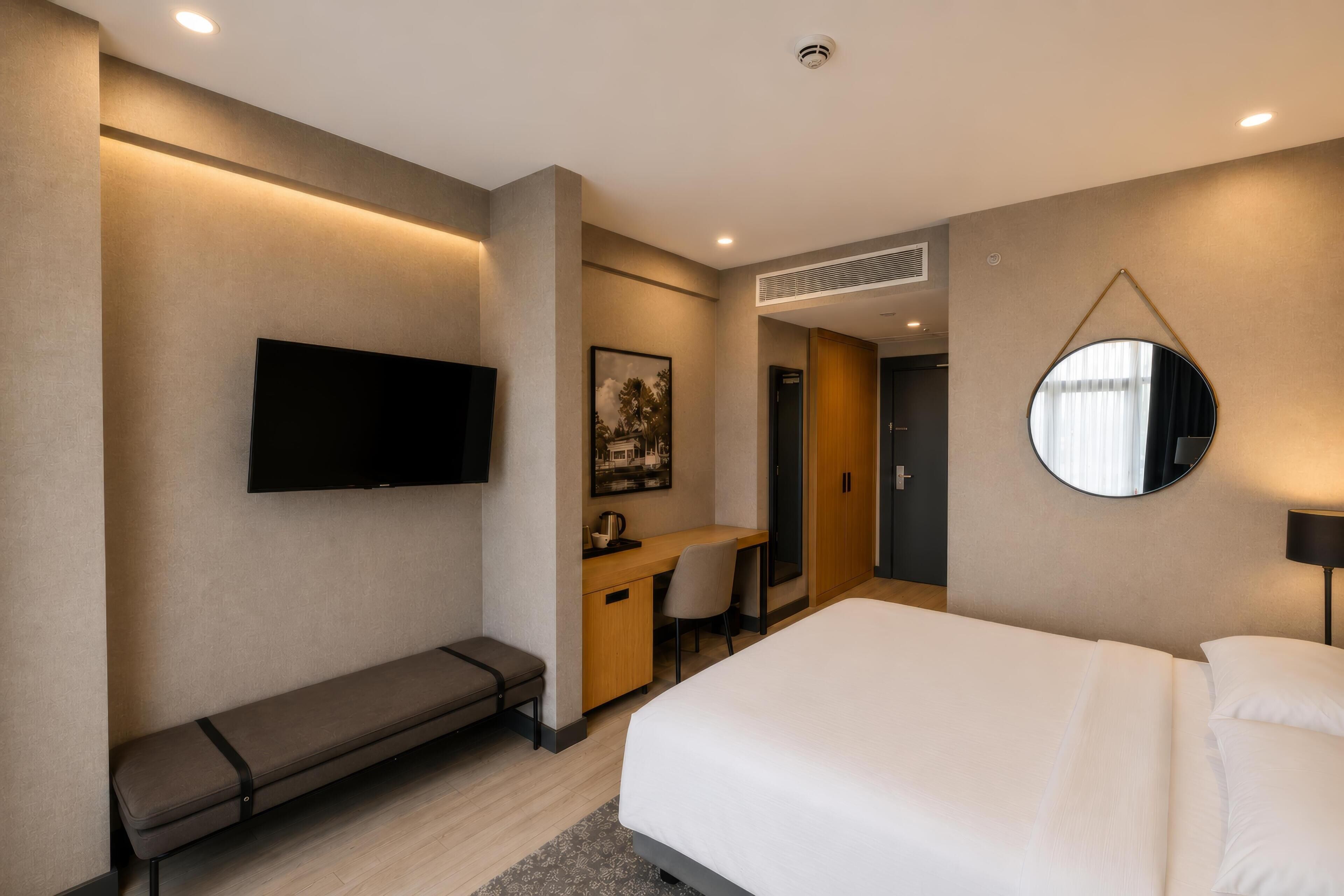 Una habitación de hotel moderna con una cama, un escritorio, un banco, un televisor de pantalla plana y un espejo redondo.