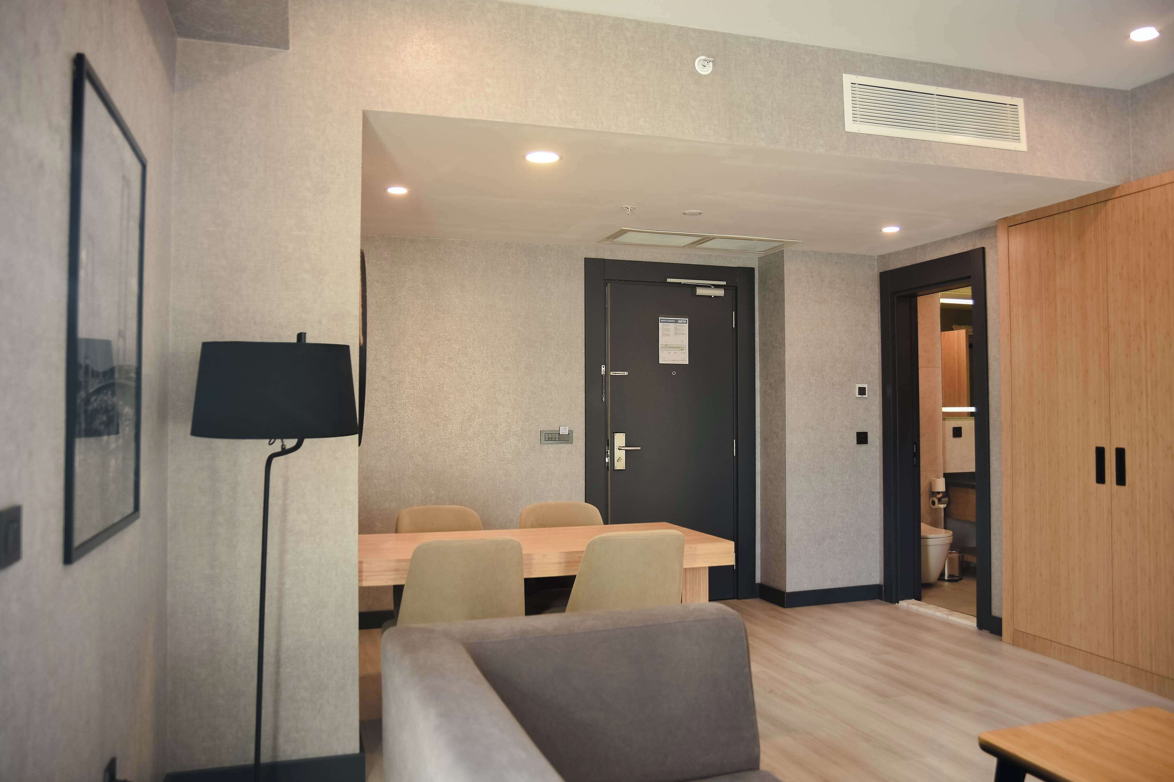 A hotel room with a wooden dining table, a sofa, a lamp, a door with a notice, and a wardrobe.