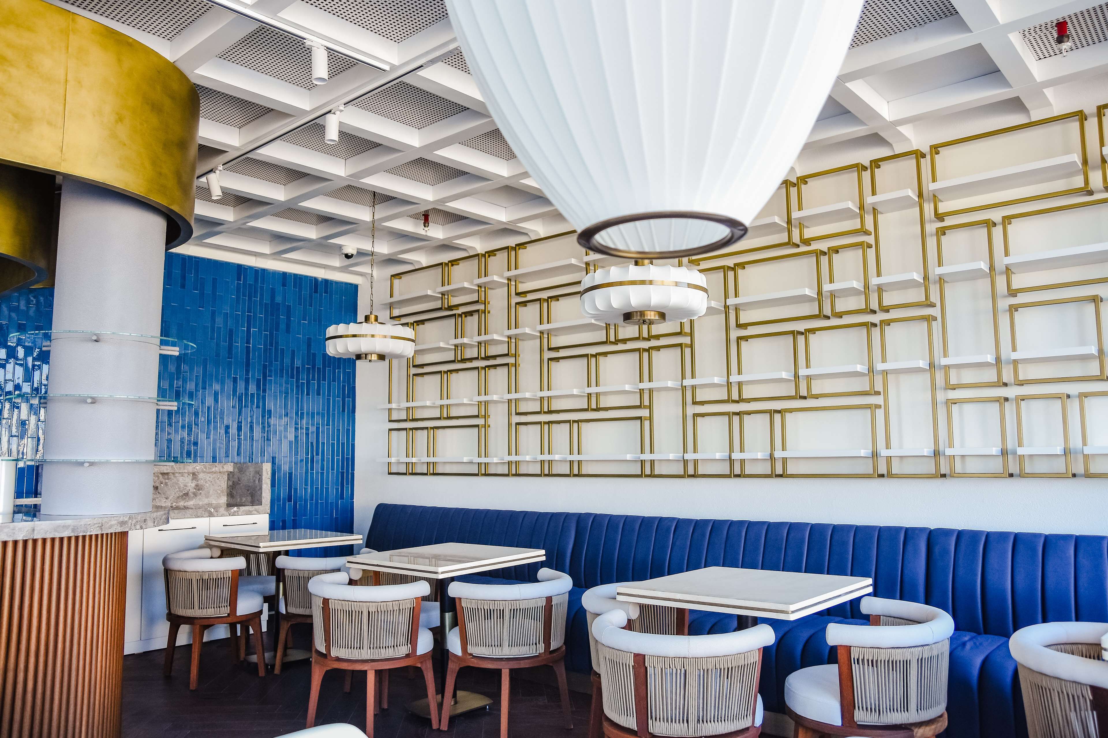 Interior de restaurante con cabinas tapizadas en azul, mesas blancas, sillas de mimbre e iluminación colgante.