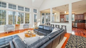A spacious living room with a large black leather sofa, a glass coffee table, and a dining area with a kitchen in the background. - Portside Villa Luxury Rental on Lake Gaston (Bracey)