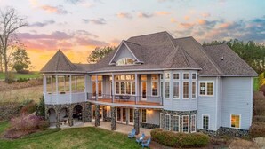 A large two-story house with a covered patio, stone pillars, and a view of the surrounding landscape. - Portside Villa Luxury Rental on Lake Gaston (Bracey)