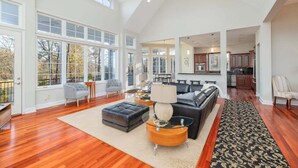 A spacious living room with a large window, a ceiling fan, and a view of the kitchen. - Portside Villa Luxury Rental on Lake Gaston (Bracey)