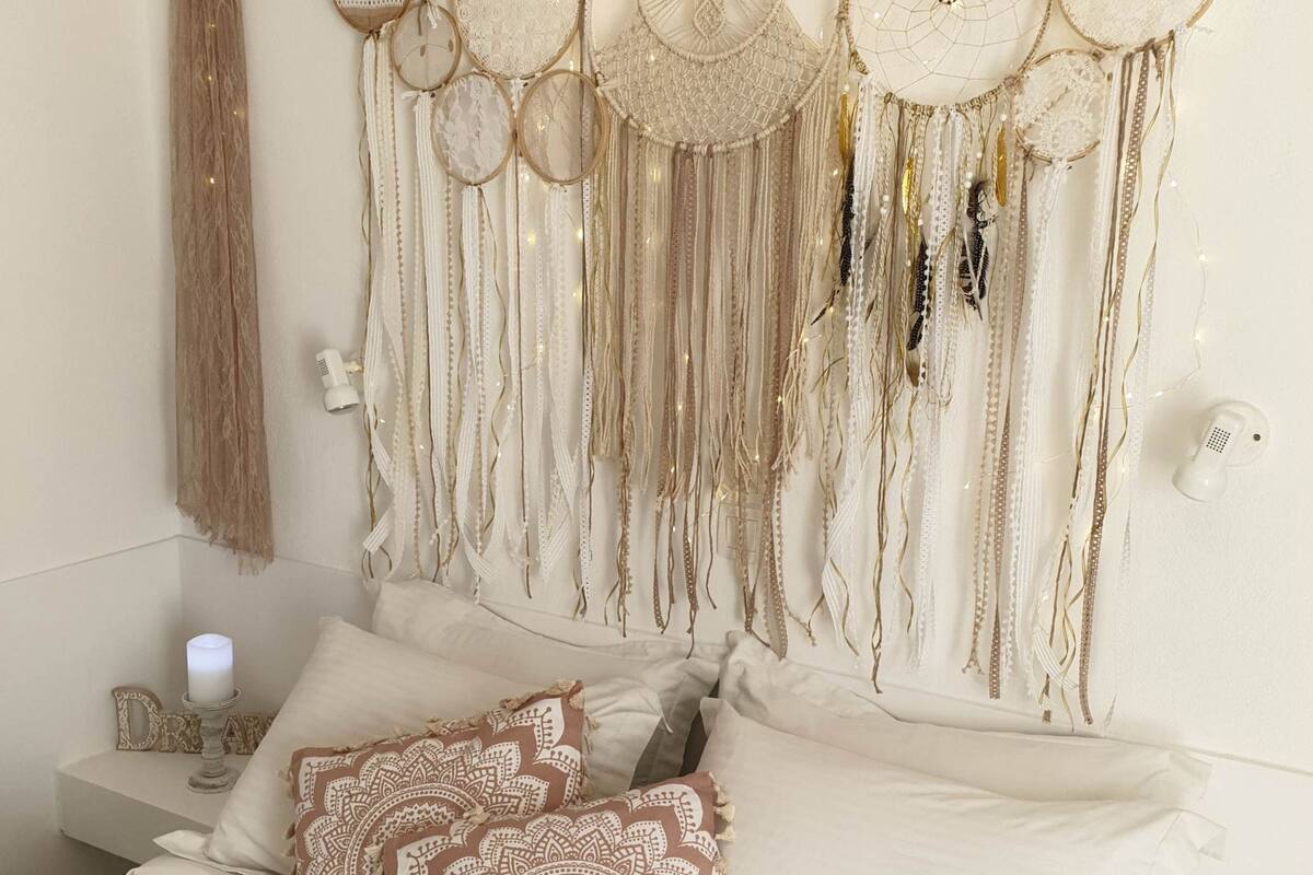A bed with white linens and patterned pillows, a wall with hanging dreamcatchers and tassels, and a bedside table with a lamp and a decorative item.