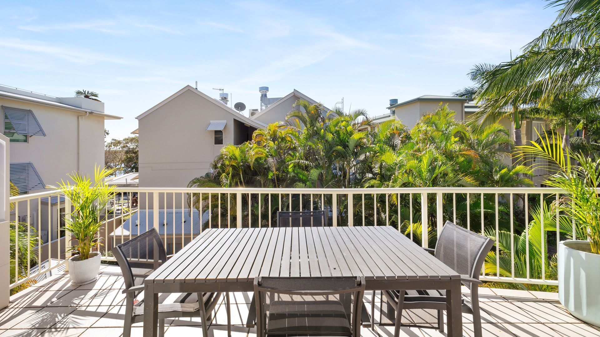 Une terrasse en bois avec une table et des chaises, entourée d’une balustrade blanche, avec des bâtiments et des plantes tropicales en arrière-plan.