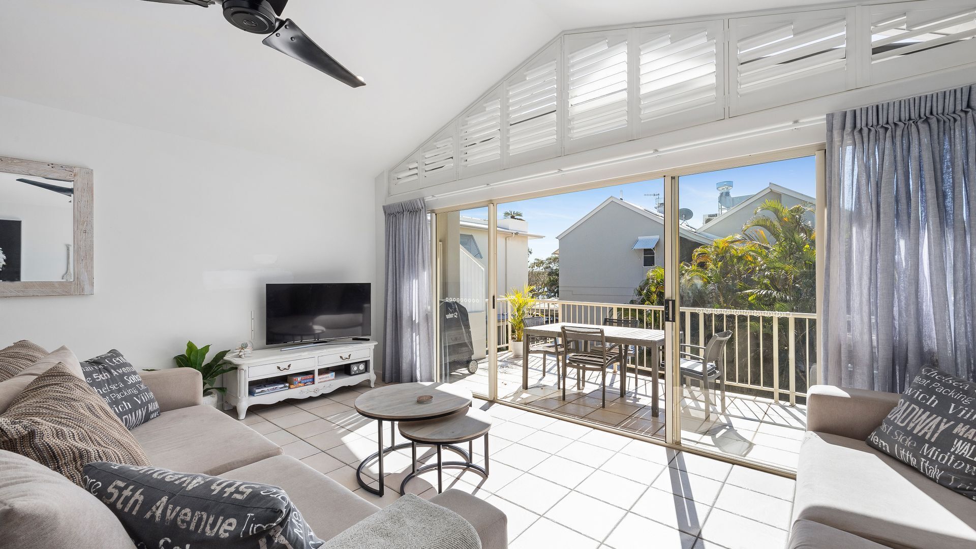 A modern living room with a flat-screen TV, a sofa, a coffee table, and a balcony with a dining area.