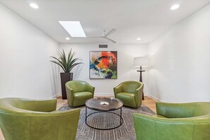 Quarto moderno com poltronas de couro verde, uma mesa de centro redonda, um abajur, uma planta em vaso e uma pintura abstrata colorida na parede.