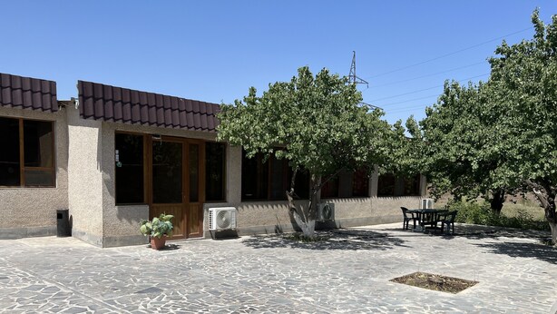 A courtyard with stone flooring, a building with large windows and a door, and a tree providing shade. - Hotel KA-EL (Parakar)