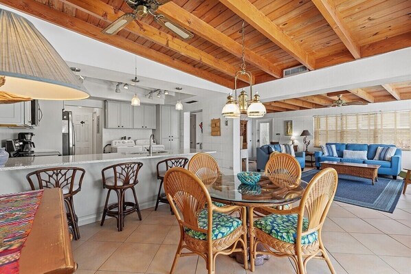 A spacious living area with a dining table surrounded by wicker chairs, a sofa, and a coffee table. A kitchen with white cabinets and a large island is visible in the background. - My Beach Key~ Beach Membership, 32' Dock, Tiki Hut (Key Colony Beach)