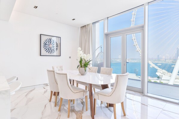 Un espace repas moderne avec une table et des chaises blanches, une plante en pot et une œuvre d’art encadrée accrochée au mur.