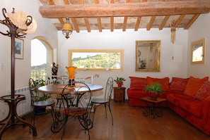 A cozy living room with a wooden ceiling, a dining area with a round table and chairs, a red sofa, and a view of greenery outside.